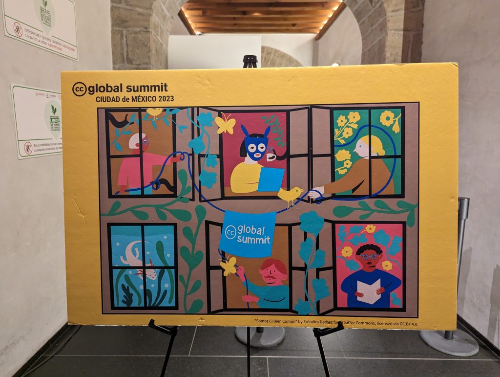 A photo of a poster for the CC Global Summit created by Erindira Derbaz. It is bright and colourful with six windows on an apartment window visible. Five people are visible through the windows and, in the bottom left-hand window an axolotl can be seen. Between the three windows in the top row is a cable with a bright blue banner hung on it and emblazoned with the CC Global Summit logo.