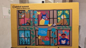 A photo of a poster for the CC Global Summit created by Erindira Derbaz. It is bright and colourful with six windows on an apartment window visible. Five people are visible through the windows and, in the bottom left-hand window an axolotl can be seen. Between the three windows in the top row is a cable with a bright blue banner hung on it and emblazoned with the CC Global Summit logo.